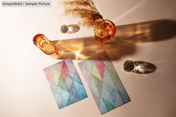 Two colorful geometric design cards on a table with glass objects casting shadows.