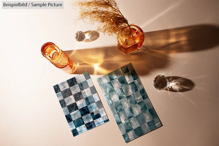 Decorative orange glasses and two checkered blue-green sheets on a beige surface with shadow play.