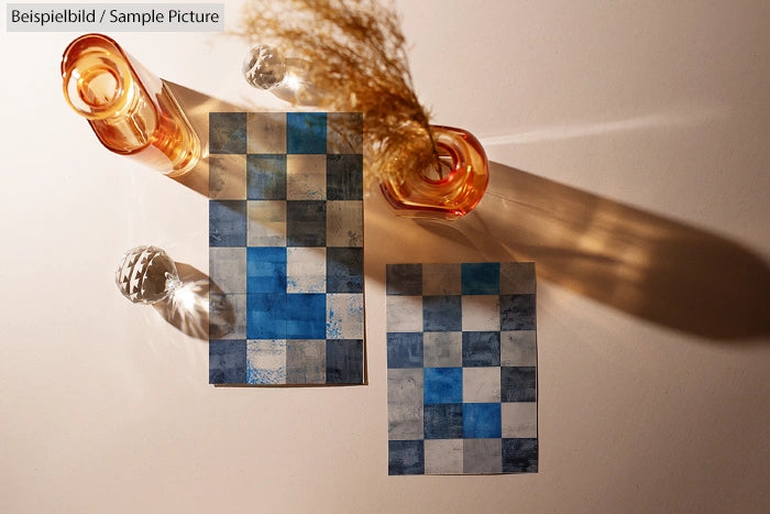 Abstract checkered art pieces with blue tones alongside orange glass vases casting shadows on a white surface.