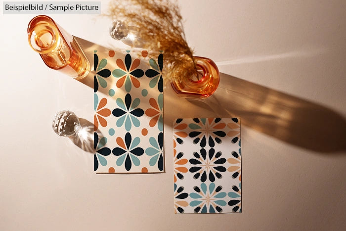 Geometric floral pattern on paper, glass bottles casting shadows, and dried plants on a beige surface.