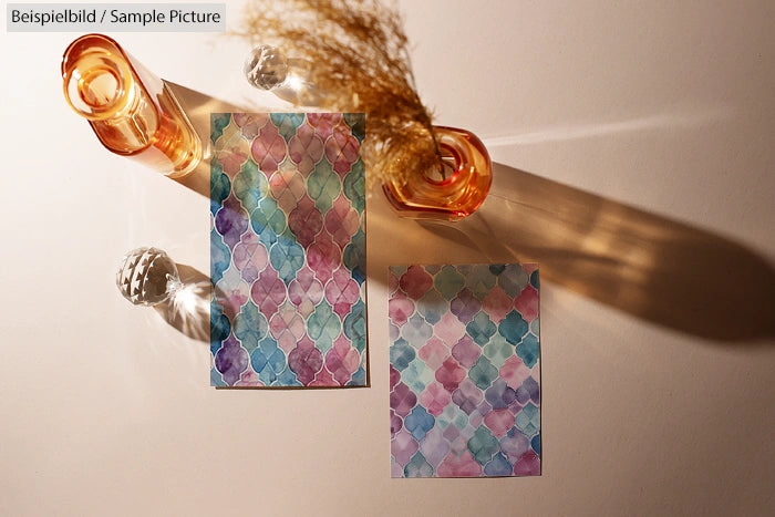 Two watercolor pattern cards with geometric designs on a sunlit table, accompanied by glass vases casting shadows.