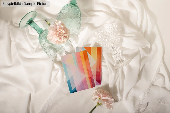 Colorful postcards with geometric patterns on white fabric, surrounded by crystal glassware and pink flowers.