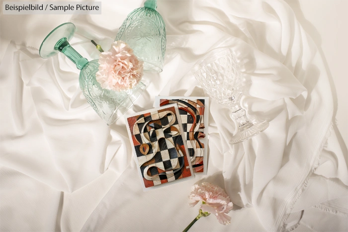 Artistic photo with two pink carnations, vintage glasses, and abstract checkerboard paintings on a white fabric background.