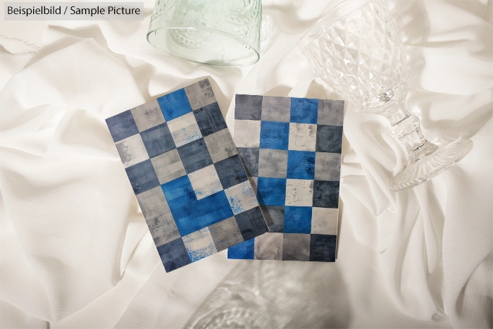 Two blue and gray checkered tiles on a white fabric backdrop with glassware.