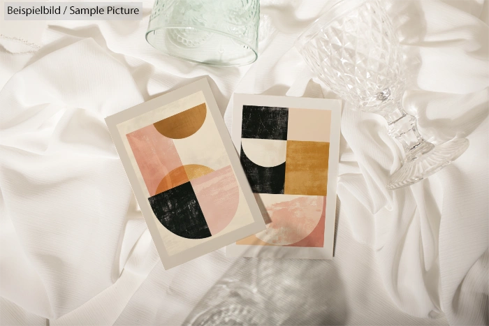 Two abstract art prints on white fabric, featuring geometric shapes in black, gold, pink, and beige tones.