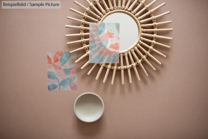 Round woven mirror with art prints of pastel leaves on a beige background, accompanied by a small empty bowl.