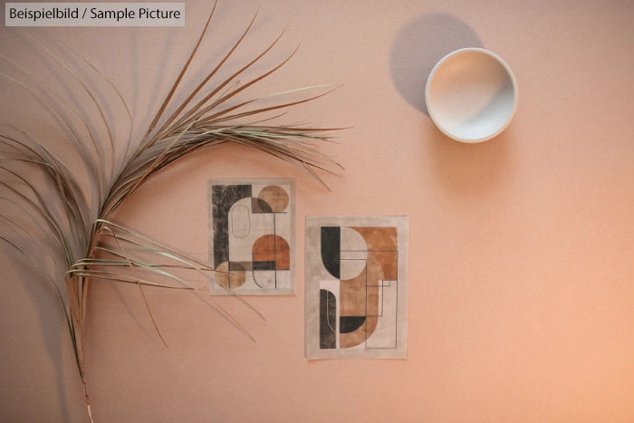 Minimalist decor with dried palm branch, two abstract art cards, and shadow of circular bowl on beige wall.