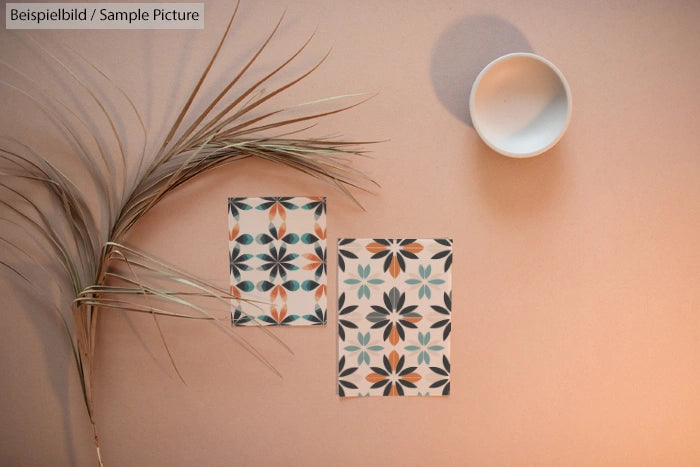 Decorative tiles with floral patterns next to palm frond and empty cup on beige surface.