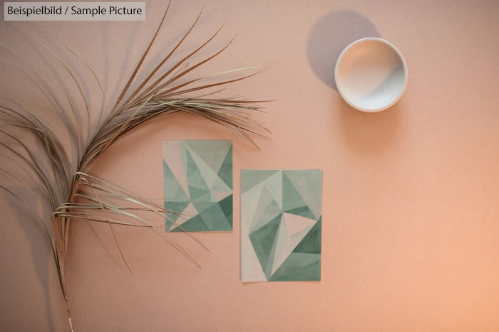 Geometric art prints on a beige surface with dried palm leaf and white bowl casting shadow.