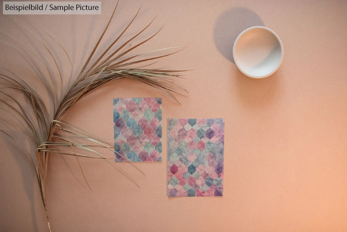 Two colorful geometric pattern cards on peach surface with ornamental grass and empty bowl.