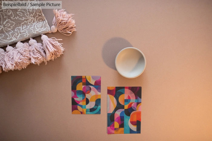 Two colorful geometric abstract prints on a table with a tassel cushion and an empty ceramic bowl.