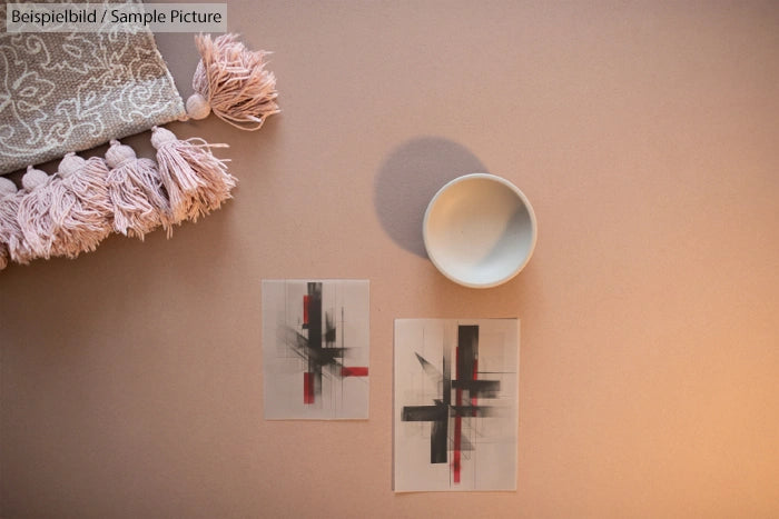 Minimalist still life with two abstract sketches, a white bowl, and a fringed textile on a beige background.