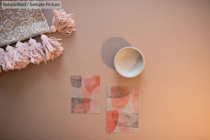 Overhead view of two abstract pink and gray art pieces next to a small white bowl on a beige surface with a tassel cloth.