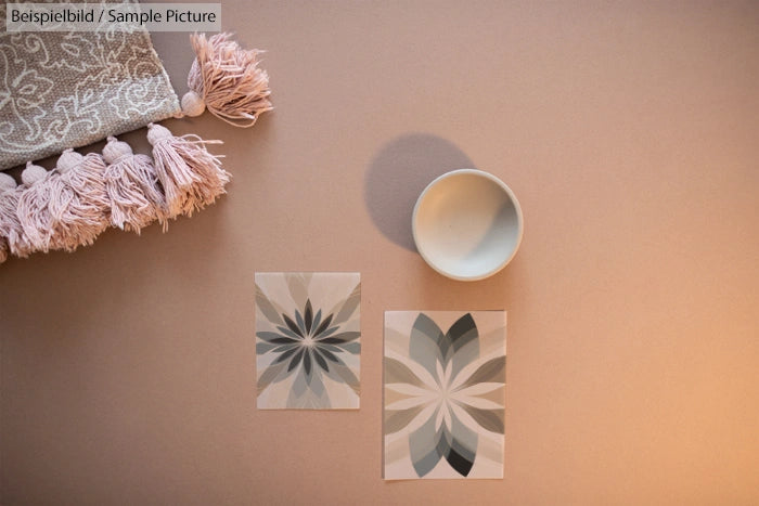 Flat lay with tasseled textile and geometric floral cards on a beige background, next to a small round bowl.