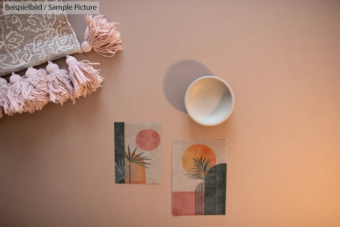 Minimalist art prints with pink and orange hues, alongside a white bowl and tasseled fabric on a peach surface.