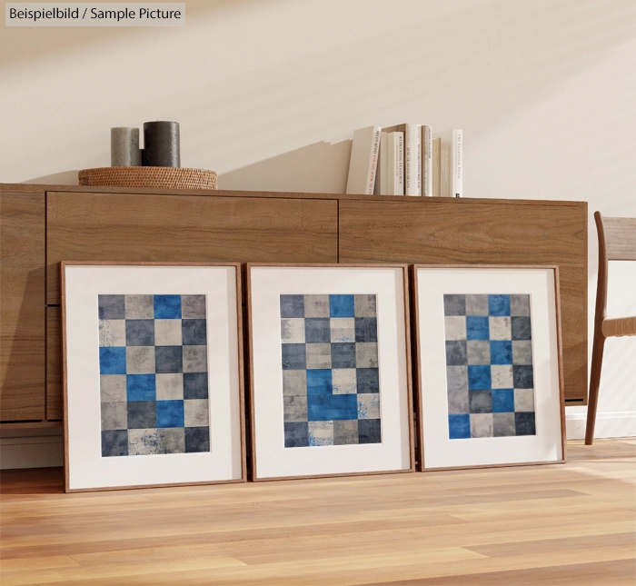 Three abstract paintings with blue, gray, and white checkered patterns on a wooden floor against a light wooden cabinet.