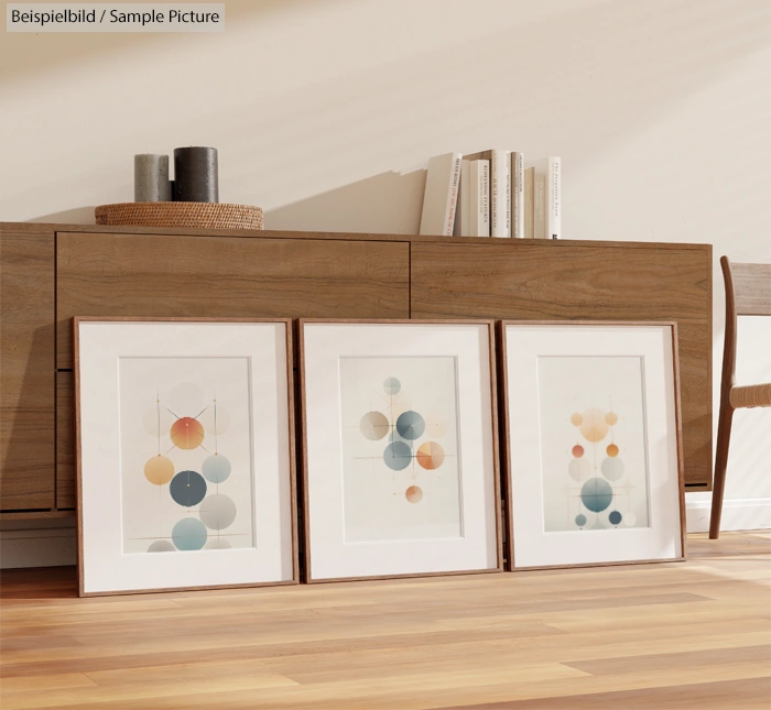 Three framed abstract art prints with geometric shapes on floor against a wooden sideboard and neutral wall.