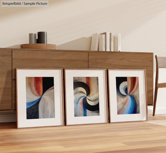 Three abstract paintings with swirling patterns, framed and leaning against a wooden cabinet in a modern interior.