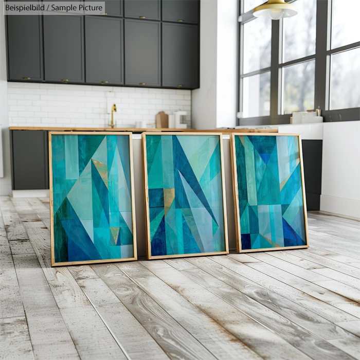 Three abstract blue and green geometric paintings on the floor of a modern kitchen with large windows and gray cabinets.