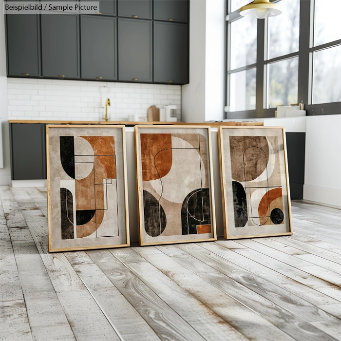 Three abstract art paintings in frames on a wooden floor with a modern kitchen and large windows in the background.