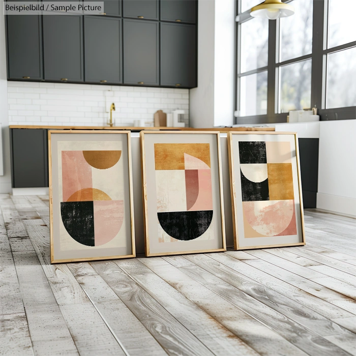 Three framed abstract geometric artworks on a wooden floor in a kitchen with sleek black cabinets and white tiles.