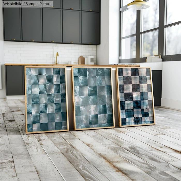 Three framed mosaic art pieces with blue and gray tones displayed on wooden floor in modern kitchen setting.