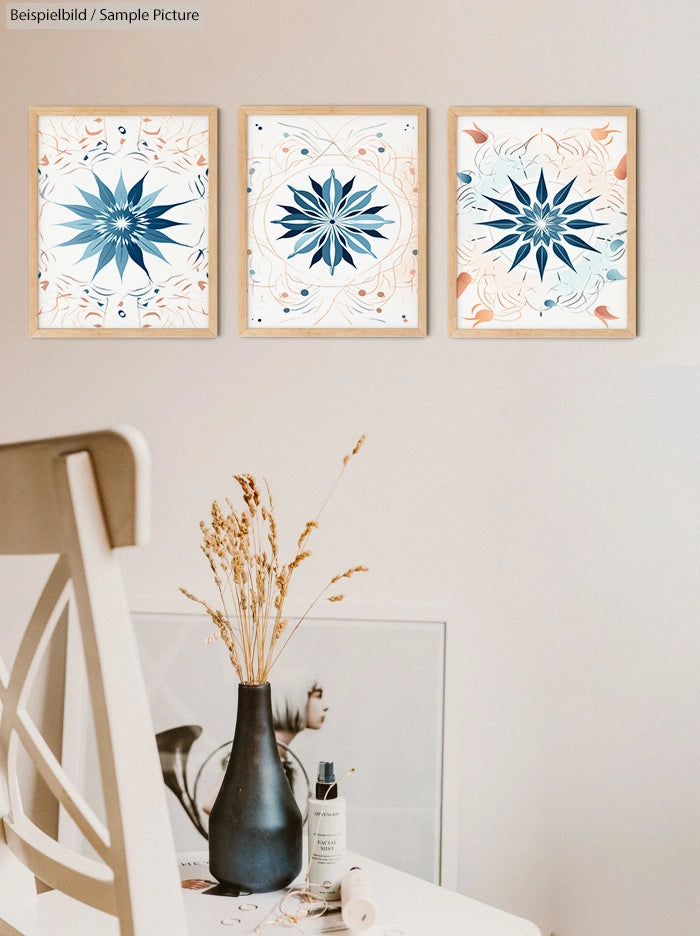 Three framed geometric artworks on a wall above a table with dried flowers and cosmetic bottle.