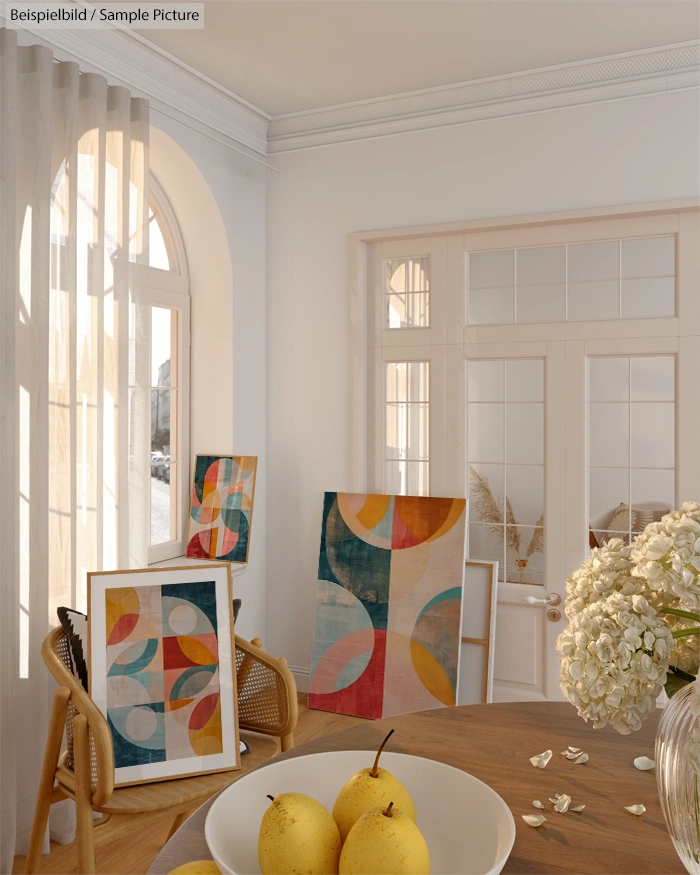 Bright room with abstract art on canvases, wooden round table, pears in bowl, and hydrangeas, near arched windows.