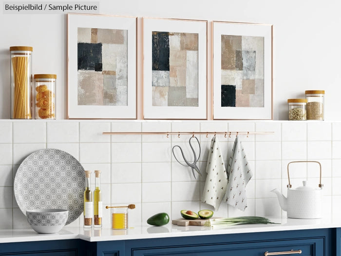 Modern kitchen with abstract framed art, blue cabinets, avocado, decorative plates, and a white kettle on tiled backsplash.