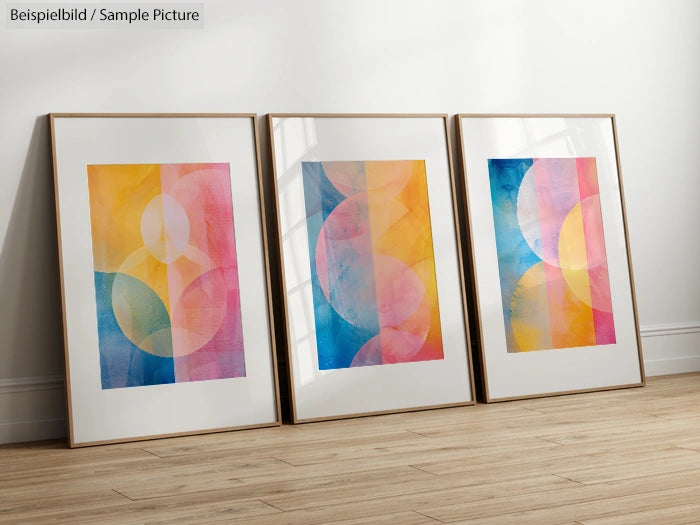 Three framed abstract art pieces with geometric patterns in shades of pink, blue, and yellow, displayed on a wooden floor.